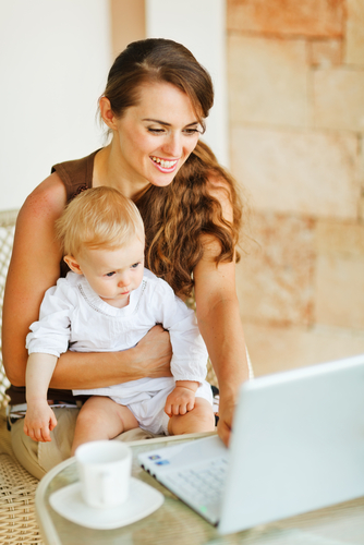 A woman sitting at a table holding a baby while looking at a laptop.