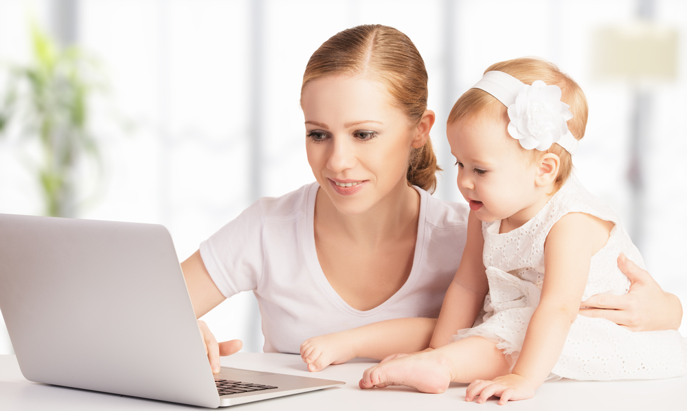 A baby sitting on a table while a woman looks at  laptop.