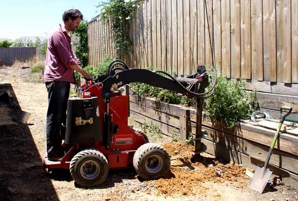 A person operating a large power tool, possibly an earth auger, on a construction site with other tools nearby.